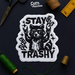 May include: Embroidered patch with a black and white raccoon wearing sunglasses, giving a rock and roll hand gesture. The text "STAY TRASHY" is above and below the raccoon. The patch has a white border and is on a dark blue surface.