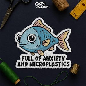 May include: Embroidered patch featuring a cartoon fish in shades of blue, yellow, and pink. The patch includes the text "FULL OF ANXIETY AND MICROPLASTICS" in white. The patch is surrounded by sewing supplies.