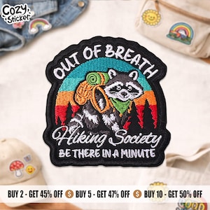 May include: Embroidered patch with a raccoon wearing a green bandana and carrying a backpack, with the text "OUT OF BREATH Hiking Society BE THERE IN A MINUTE". The patch has a black border and a colorful mountain scene.