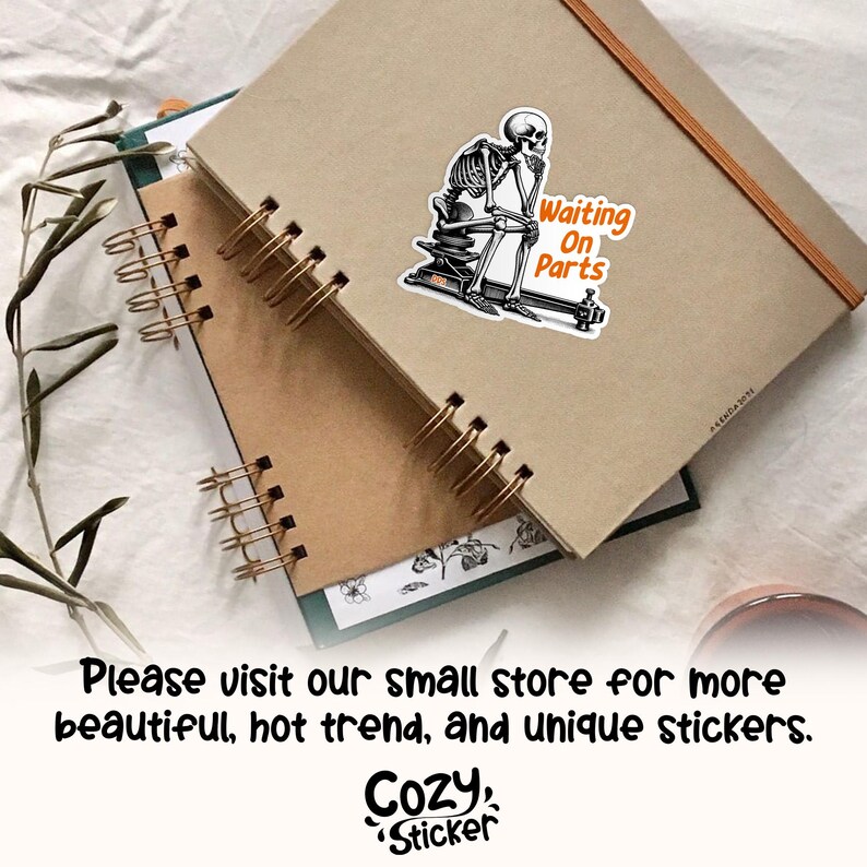 May include: A sticker featuring a skeleton sitting on a machine, with the text "Waiting On Parts" in orange. The sticker is placed on a brown notebook. The image also includes a spiral-bound notebook and the text "Cozy Sticker".