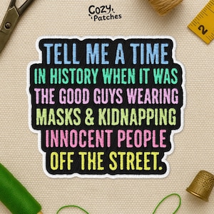 May include: Embroidered patch with the text "TELL ME A TIME IN HISTORY WHEN IT WAS THE GOOD GUYS WEARING MASKS & KIDNAPPING INNOCENT PEOPLE OFF THE STREET." The patch has a black background with colorful text and a white border. The patch is on a beige fabric.