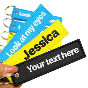 May include: A collection of rectangular keychains in blue, yellow, and black. The blue keychain reads "Look at my eyes", the yellow keychain says "Jessica", and the black keychain says "Your text here". Each keychain has a silver metal ring.