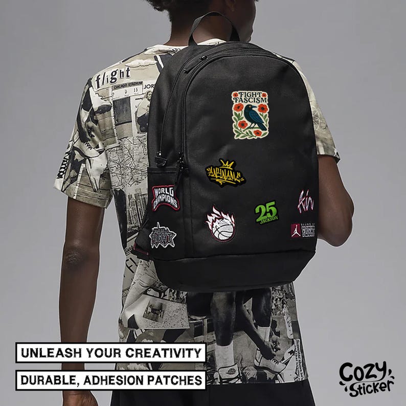 Puede incluir: Mochila negra adornada con varios parches coloridos. Los parches incluyen textos como "FIGHT FASCISM", "WORLD CHAMPIONS" y un gr&aacute;fico de baloncesto. La mochila tambi&eacute;n presenta el texto "UNLEASH YOUR CREATIVITY" y "DURABLE, ADHESION PATCHES".
