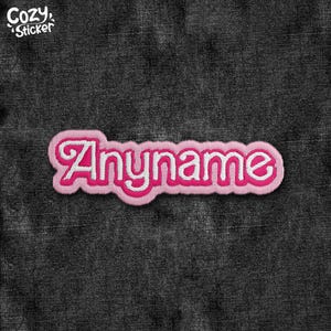 May include: A pink and white embroidered patch with the word "Anyname" in a cursive font. The patch has a pink border and is set against a dark gray denim background. The text "Cozy Sticker" is in the upper left corner.