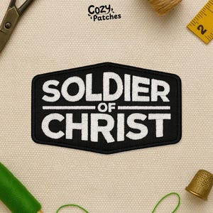 May include: Black embroidered patch with the words "SOLDIER OF CHRIST" in white, stylized lettering. The patch has a black border and is set against a neutral background. The patch is likely for clothing or accessories.