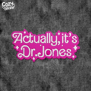 May include: A pink embroidered patch with the text "Actually, it's Dr. Jones" in white script. The patch has a decorative border and is set against a dark gray denim background. The Cozy Sticker logo is in the upper left corner.