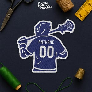 Peut inclure: Écusson brodé représentant un joueur de crosse en maillot bleu avec le nom "ANYNAME" et le numéro "00". Le patch a une bordure blanche. Le logo "Cozy Patches" est visible. Fond bleu foncé.