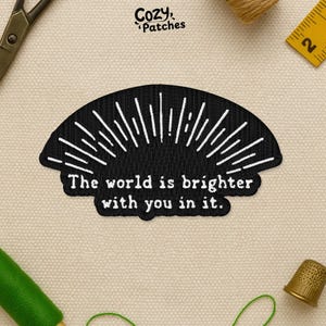 May include: Black embroidered patch in a half-circle shape with white radiating lines and the text "The world is brighter with you in it." The patch is from Cozy Patches and is surrounded by sewing tools.
