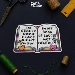 May include: Embroidered patch in the shape of an open book with the text "IN REALLY GOOD PLACE RIGHT NOW IN MY BOOK OF COURSE NOT MENTALLY". The patch has a white background with colorful accents and floral and heart designs.