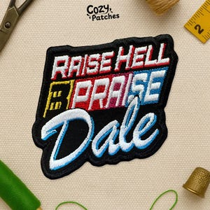 May include: Embroidered patch with the text "RAISE HELL PRAISE Dale" in white, red, blue, and yellow stitching on a black background. The patch has a black border and is surrounded by sewing supplies.