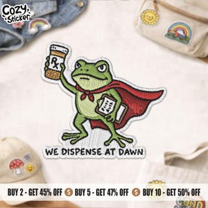 Peut inclure: Un écusson brodé représentant une grenouille verte portant une cape rouge, tenant un flacon de médicament dans une main et un presse-papier dans l'autre. Le texte WE DISPENSE AT DAWN est brodé en noir sous la grenouille sur fond blanc.