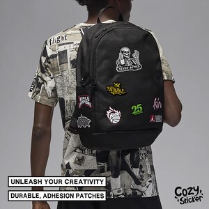 Puede incluir: Mochila negra adornada con parches coloridos, incluyendo un esqueleto, una pelota de baloncesto y texto. Los parches dicen "NEVER BETTER", "WORLD CHAMPIONS" y "25". La mochila tiene un bolsillo lateral y un asa superior. Tambi&eacute;n se ve el texto "UNLEASH YOUR CREATIVITY" y "DURABLE, ADHESION PATCHES".