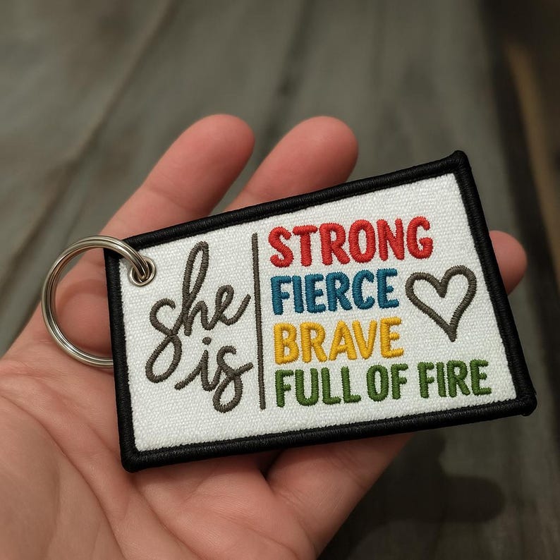 May include: Embroidered keychain with the words "she is" in a script font, and the words "STRONG FIERCE BRAVE FULL OF FIRE" in colorful block letters. The keychain has a black border and a silver key ring.