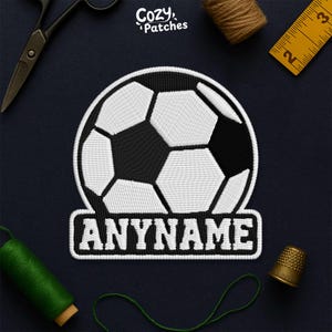 May include: Embroidered soccer ball patch with the text "ANYNAME" in a white, bold font. The patch is black and white, with a soccer ball design. The patch is surrounded by sewing tools and thread.