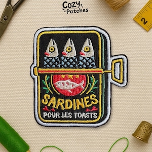 May include: Embroidered patch featuring a sardine can design. The patch has a black background with a yellow border. The can contains three sardines with red accents. Text on the patch reads "Sardines Pour Les Toasts". The patch is from Cozy Patches.