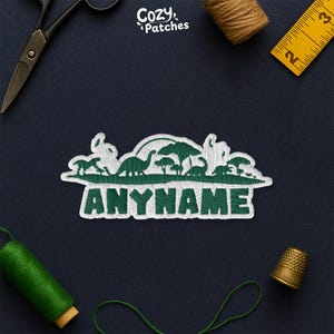 May include: Embroidered patch featuring a green dinosaur landscape with the text "ANYNAME" in green. The patch is white with green stitching. The image includes sewing tools and thread on a dark blue surface.