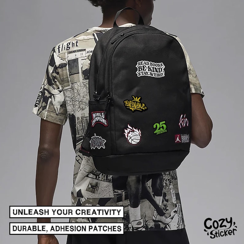 May include: Black backpack with a variety of colorful patches, including "Read Books Be Kind Stay Weird", "World Champions", "25", and a basketball with flames. The backpack is worn by a person wearing a white and black patterned shirt.