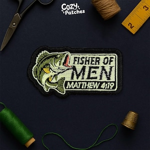 May include: Embroidered patch featuring a green and yellow fish with the text "FISHER OF MEN" and "MATTHEW 4:19" in white. The patch is black with a white border. The image also includes sewing tools and thread.