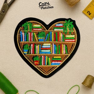 May include: Heart-shaped patch with a bookshelf design, displaying colourful books and potted plants. The patch has a brown background with a black border and the text "Cozy Patches" in the upper left corner.