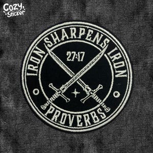 May include: Black and white embroidered patch with the text "IRON SHARPENS IRON PROVERBS 27:17" surrounding two crossed swords. The patch is circular and has a white border. The patch is on a gray denim background.