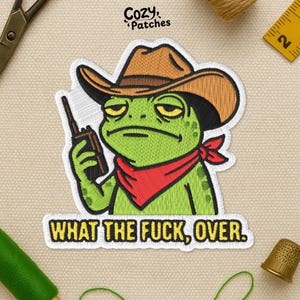 May include: Embroidered patch featuring a green frog wearing a cowboy hat and red bandana, holding a walkie-talkie. The patch has the text "WHAT THE FUCK, OVER." in a bold font. The patch is from Cozy Patches.