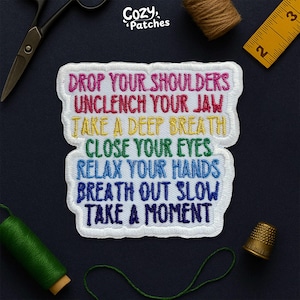 May include: A white embroidered patch with the text "DROP YOUR SHOULDERS, UNCLENCH YOUR JAW, TAKE A DEEP BREATH, CLOSE YOUR EYES, RELAX YOUR HANDS, BREATH OUT SLOW, TAKE A MOMENT" in rainbow colors. The patch is surrounded by sewing supplies.