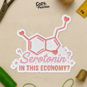 May include: Embroidered patch featuring a serotonin molecule design in pink and white, with the text "Serotonin? IN THIS ECONOMY?". The patch has a heart-shaped detail at the top and is surrounded by a white border. The brand name "Cozy Patches" is at the top.