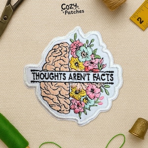 May include: Embroidered patch featuring a brain design on the left and colorful flowers on the right. The text "THOUGHTS AREN'T FACTS" is embroidered in black on a white banner. The patch has a white border and the brand name "Cozy Patches" is in the top right.