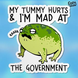 May include: A green frog with a grumpy expression and a mustache says "My tummy hurts & I'm mad at the government." The frog is on a white sticker with a "Cozy Sticker" logo in the top right corner.