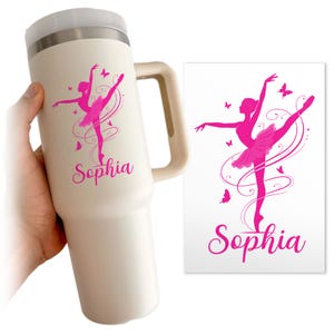 May include: A cream-colored travel mug with a handle, featuring a pink ballerina design with butterflies and the name "Sophia." A matching pink ballerina design is on a white card. The mug is designed to hold a large quantity of liquid.