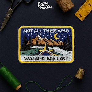 May include: Embroidered patch with a mountain and night sky design. The patch features the text "NOT ALL THOSE WHO WANDER ARE LOST" in white. The patch is rectangular with a yellow border and a blue background.