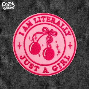 May include: A pink circular patch with the text "I AM LITERALLY JUST A GIRL" in a circle around a pair of cherries with a bow. The patch has a light pink background and a darker pink border and design.