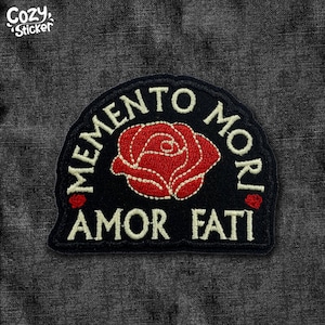 May include: Black patch with embroidered text and a red rose. The patch reads "MEMENTO MORI" in a semi-circle above "AMOR FATI". The rose is in the center, with white stitching. The patch is on a dark gray textured background.