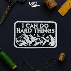 May include: A black and white embroidered patch featuring the phrase "I CAN DO HARD THINGS" above a mountain range design. The patch has a white border. The brand name "Cozy Patches" is in the top left corner.