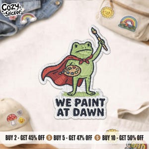 May include: An embroidered patch featuring a green frog wearing a red cape, holding a paintbrush in one hand and a paint palette in the other. Below the frog, the text reads WE PAINT AT DAWN in bold black letters. The patch has a white border.