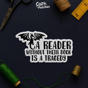 May include: Embroidered patch with the text "A READER WITHOUT THEIR BOOK IS A TRAGEDY" and a dragon design. The patch is white with black accents, set against a dark blue background. Sewing tools and thread spools surround the patch.