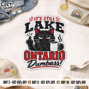 Peut inclure: Un écusson brodé représentant un chat noir tenant une tasse de café blanche et faisant un doigt d'honneur. Le texte indique IT'S STILL LAKE ONTARIO DUMBASS! Le motif utilise du fil noir, blanc et rouge sur un fond blanc avec une bordure cousue.