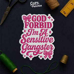 May include: Embroidered patch with the text "GOD FORBID I'm A Sensitive Gangster" in pink and white. The patch features bow accents and a decorative border. The patch is surrounded by sewing tools and supplies.