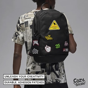 Pode incluir: Mochila preta adornada com v&aacute;rios patches coloridos, incluindo um sinal de aviso amarelo com um p&aacute;ssaro, uma bola de basquete e o n&uacute;mero 25. A mochila tem um bolso lateral e &eacute; usada por uma pessoa vestindo uma camiseta gr&aacute;fica.