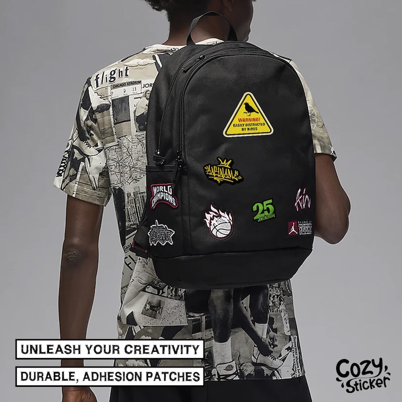 Pode incluir: Mochila preta adornada com v&aacute;rios patches coloridos, incluindo um sinal de aviso amarelo com um p&aacute;ssaro, uma bola de basquete e o n&uacute;mero 25. A mochila tem um bolso lateral e &eacute; usada por uma pessoa vestindo uma camiseta gr&aacute;fica.