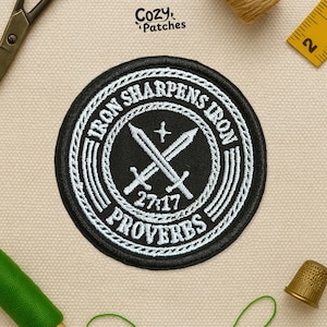 May include: A black and white embroidered patch with the text "IRON SHARPENS IRON PROVERBS 27:17" encircling two crossed swords. The patch has a decorative border and the brand name "Cozy Patches" is in the upper left corner.
