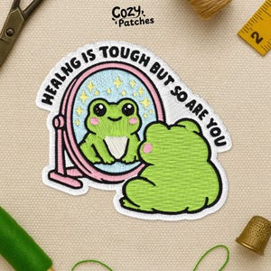 May include: Embroidered patch featuring a green frog looking in a pink mirror. The mirror reflects the frog with a light blue background and yellow stars. Text around the mirror reads "HEALING IS TOUGH BUT SO ARE YOU". The patch is from Cozy Patches.