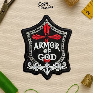 May include: Black and white embroidered patch with the text "ARMOR OF GOD" and a red cross design. The patch has a decorative border and is likely for clothing or accessories. The brand name "Cozy Patches" is at the top.