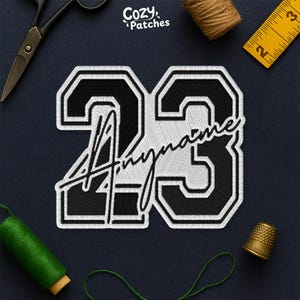 May include: Embroidered patch with the number "23" in black with a white border, and the word "Anyname" in script. The patch has a varsity style design. The brand name "Cozy Patches" is in the upper right corner.
