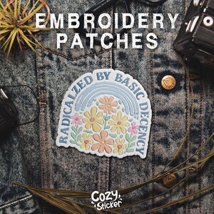 May include: Embroidered patch with a floral design and the text "RADICALIZED BY BASIC DECENCY" in a rainbow arc. The patch features pastel colours and is set against a denim jacket background. The word "Cozy Sticker" is also visible.
