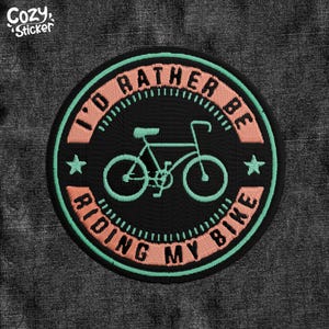May include: Embroidered patch with the text "I'D RATHER BE RIDING MY BIKE" in a circular design. The patch features a bicycle graphic in the center, with a teal outline and a black background. The text is in a peach colour, with a teal border.