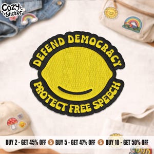 May include: Embroidered patch featuring a yellow lemon design with the text "DEFEND DEMOCRACY PROTECT FREE SPEECH" in black. The patch is surrounded by other patches and accessories.