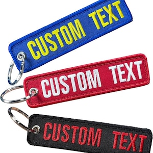 May include: Three rectangular keychains with the text "CUSTOM TEXT". The top keychain is blue with yellow text, the middle one is red with white text, and the bottom one is black with red text. Each keychain has a silver key ring.
