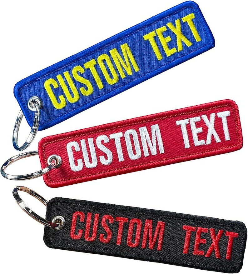 May include: Three rectangular keychains with the text "CUSTOM TEXT". The top keychain is blue with yellow text, the middle one is red with white text, and the bottom one is black with red text. Each keychain has a silver key ring.