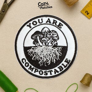 May include: A circular patch with the text "YOU ARE COMPOSTABLE" in a black and white design. The patch features a cluster of mushrooms with detailed caps and roots. The patch is surrounded by a white border.